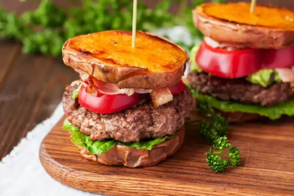 Pyszny burger na talerzu z soczystą wołowiną, chrupiącym bekonem, pomidorem i awokado, podany na słodkich ziemniakach.