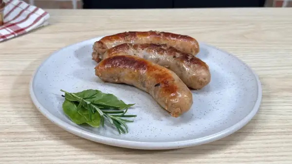 Tre salsicce dorate e succulente, appena cotte in padella, riposano su un piatto con rosmarino e alloro.