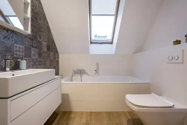 Przytulna łazienka na poddaszu z wanną, umywalką i toaletą. Dachowe okno wpuszcza naturalne światło.