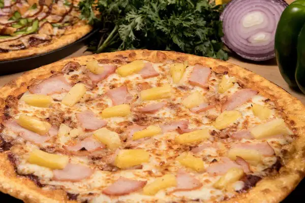 Pizza hawajska składniki: kawałki ananasa i szynki na serze. W tle inna pizza i warzywa.