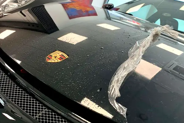 Czarna maska Porsche z logo, na której widać krople wody i fragment folii. Trwa klejenie folii na sucho.