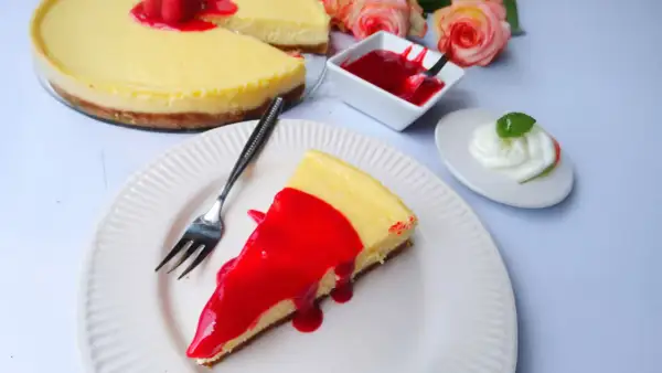 Ein Stück New York Cheesecake mit roter Fruchtsauce und Himbeeren, daneben eine kleine Schüssel Sauce und ein Klecks Sahne.