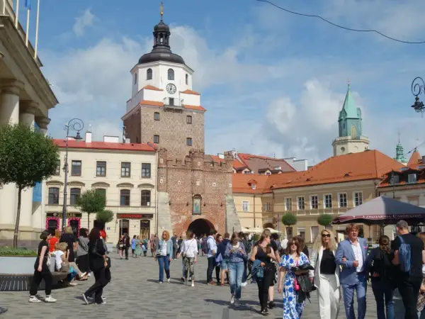 Ludzie spacerują po brukowanym placu litewskim, podziwiając zabytkową wieżę z zegarem i zieloną iglicę w tle.