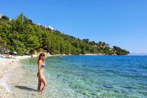 Kobieta w stroju kąpielowym i kapeluszu stoi w krystalicznie czystej wodzie na Makarska beach. W tle zielone wzgórza i błękitne niebo.