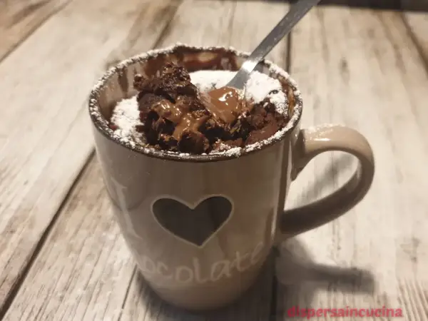 Un delizioso mug cake al cioccolato, appena sfornato, con cuore e scritta "chocolate" sulla tazza, spolverato di zucchero a velo.