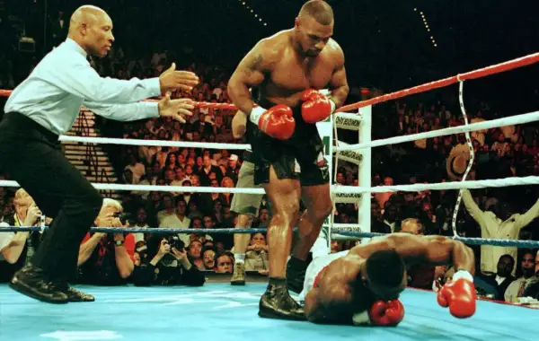 Mike Tyson stands over a fallen opponent, his powerful physique and impressive mike tyson arm length evident. The referee signals the end of the match.
