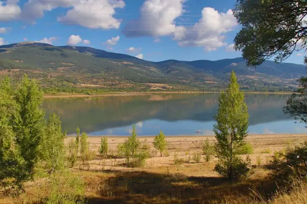 El embalse de Pinilla, con montañas verdes al fondo y un cielo azul con nubes blancas. Reflejos en el agua serena.