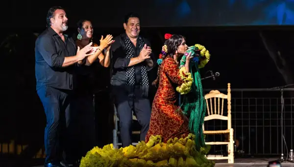 Una bailaora de flamenco andalusi, con un vestido rojo y amarillo, besa un mantón verde. A su lado, tres músicos aplauden.