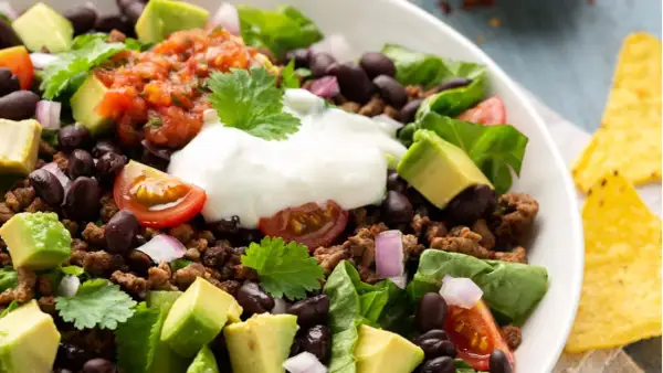 Ein bunter Taco Salat mit Hackfleisch, schwarzen Bohnen, Avocado, Tomaten, Zwiebeln und einem Klecks Sauerrahm. Perfekt für einen leichten Genuss.