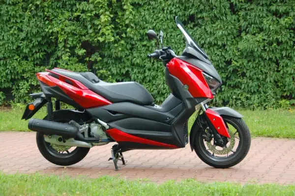 Czerwony skuter Yamaha XMAX 125, gotowy do odblokowania pełnej mocy, zaparkowany na tle zielonego żywopłotu.