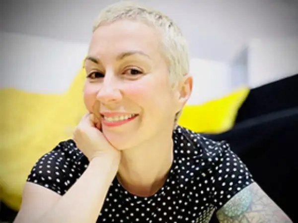 Laura Veltz, a blonde woman with short hair, smiles and rests her chin on her hand. She wears a black polka dot shirt.