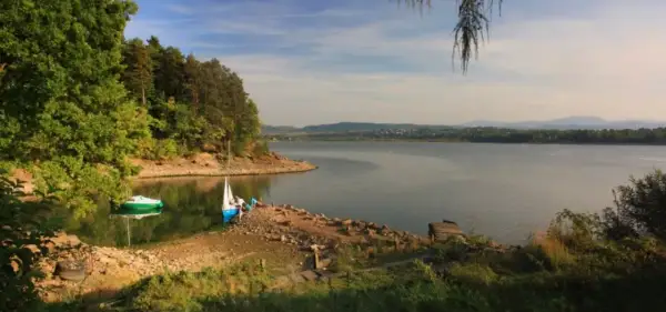 Spokojne jezioro żywieckie z łódkami przy brzegu, otoczone zielonymi drzewami i wzgórzami w oddali.