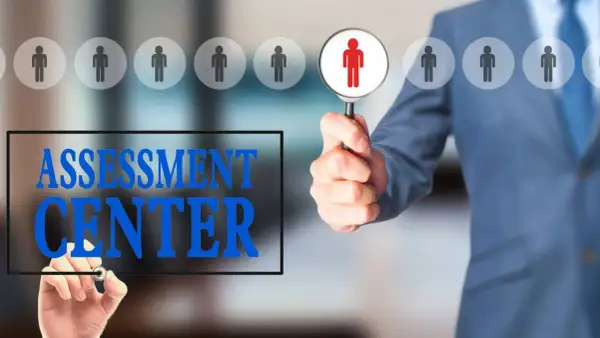 Uomo in giacca blu esamina candidati con lente d'ingrandimento. Testo "ASSESSMENT CENTER" scritto su schermo.