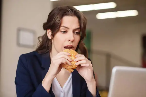 Une femme mange un hamburger à son bureau, profitant d'une pause déjeuner rapide.
