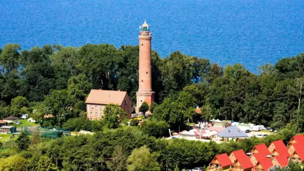 Latarnia morska Sarbinowo góruje nad zielonymi drzewami, z błękitnym morzem w tle. Obok niej budynki i czerwone dachy domków letniskowych.