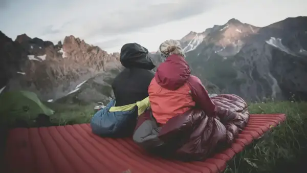 Dos personas sentadas en un colchón inflable, envueltas en sacos de dormir, disfrutan de un paisaje montañoso. El material vivac les permite acampar cómodamente.