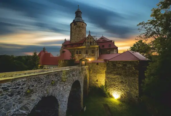 Zamek Czocha, gdzie jest, w blasku zachodzącego słońca i nocnych świateł, z kamiennym mostem i wieżą.