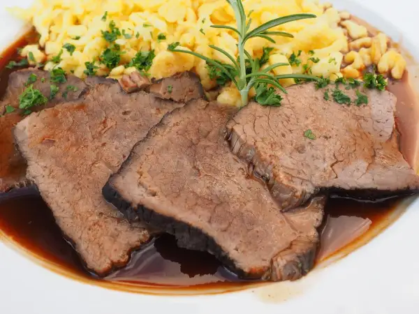 Fertig eingelegter Sauerbraten zubereiten: Zarte Scheiben Rindfleisch in dunkler Sauce mit Spätzle und Rosmarin garniert.