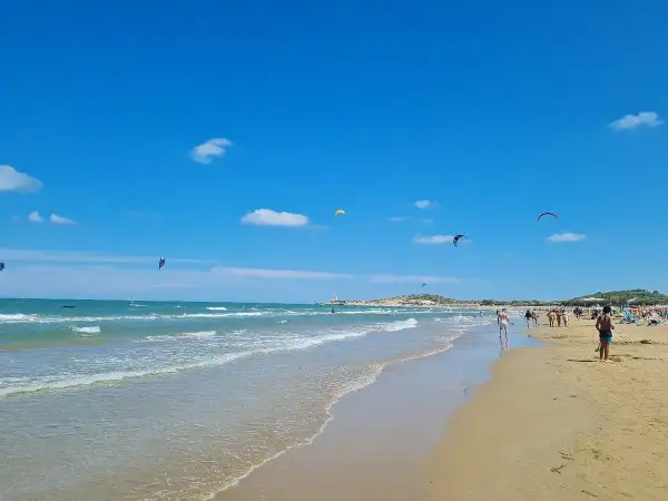 Spiaggia di Scialmarino con kitesurfer che danzano nel cielo azzurro e bagnanti che passeggiano sulla sabbia dorata.