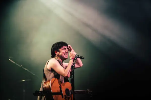 Declan McKenna passionately sings into a microphone, guitar in hand, bathed in stage lights. A glimpse of his setlist is visible.
