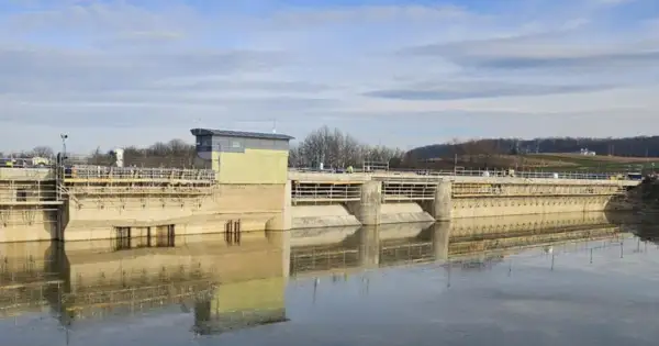 Betonowa zapora w Sieniawie w budowie, z widocznym budynkiem sterowni i rusztowaniami. Woda odbija konstrukcję.