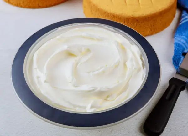 Puszysta bita śmietana z mascarpone w misce, gotowa do dekorowania tortu. Obok leżą biszkopty i szpatułka.