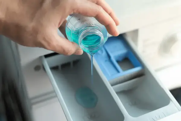 Mano versa liquido blu nel cassetto della lavatrice, mostrando dove mettere il Calgon per un bucato impeccabile.