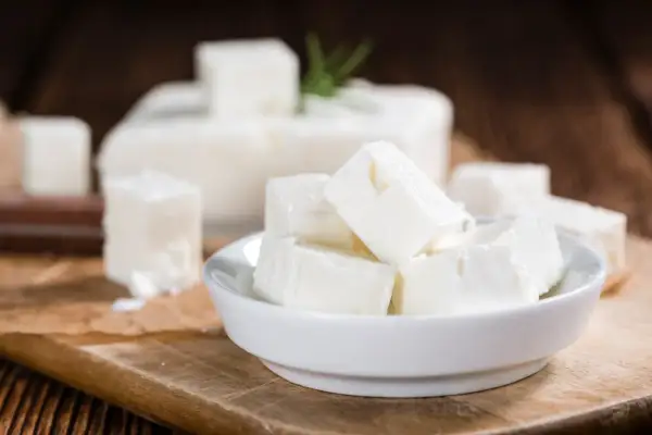 Cubos de queso de leche cruda en un cuenco blanco, con un bloque de queso y una ramita de romero al fondo.