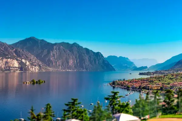 Un incantevole panorama per le tue vacanze sul Lago di Garda: montagne maestose, un'isola verde e un borgo pittoresco con barche a vela.