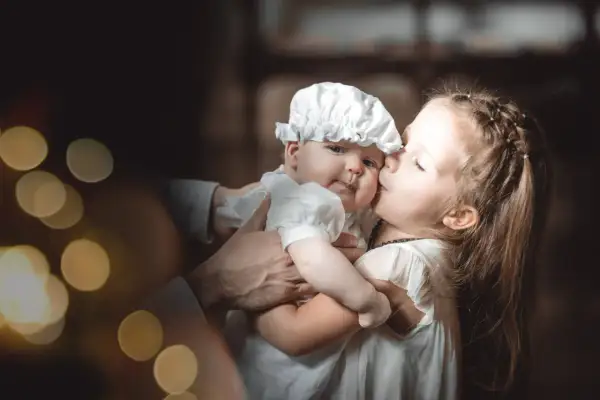 Une sœur embrasse tendrement son bébé, habillé pour baptiser son enfant. Lumières douces et ambiance chaleureuse.
