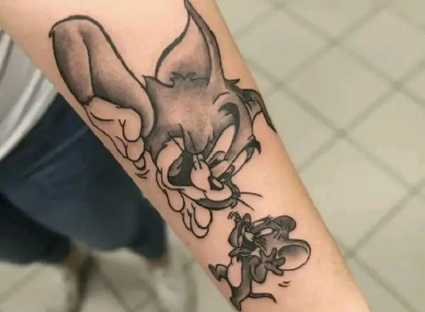 Czarno-biały tatuaż tom i jerry na przedramieniu. Kot Tom próbuje złapać myszkę Jerry, która ucieka.