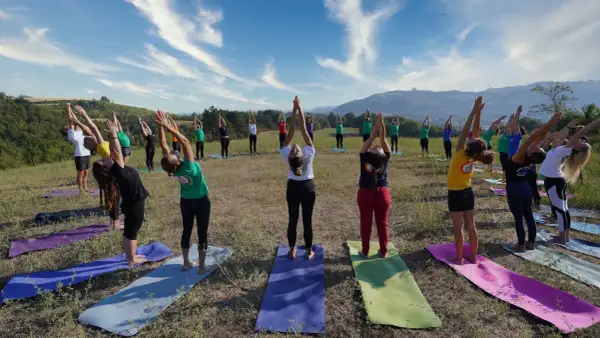 Gruppo di persone in posa urdhva hastasana su tappetini da yoga in un prato verde, con cielo azzurro e nuvole bianche.
