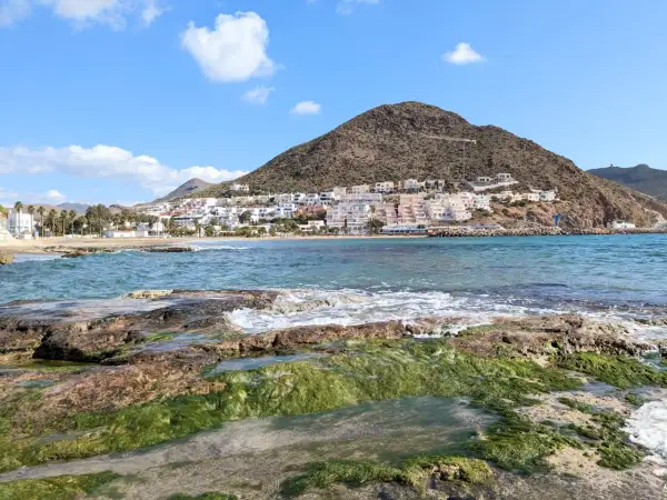 Costa de Cabo de Gata en octubre, con rocas cubiertas de algas verdes y el mar azul. Al fondo, el pueblo blanco se alza en la ladera de una montaña.