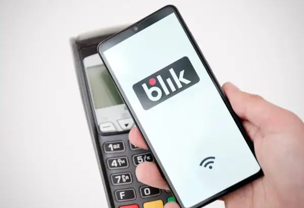 Dłoń trzyma smartfon z logo Blik nad terminalem płatniczym. Szybki przelew na telefon bez obaw o limit.