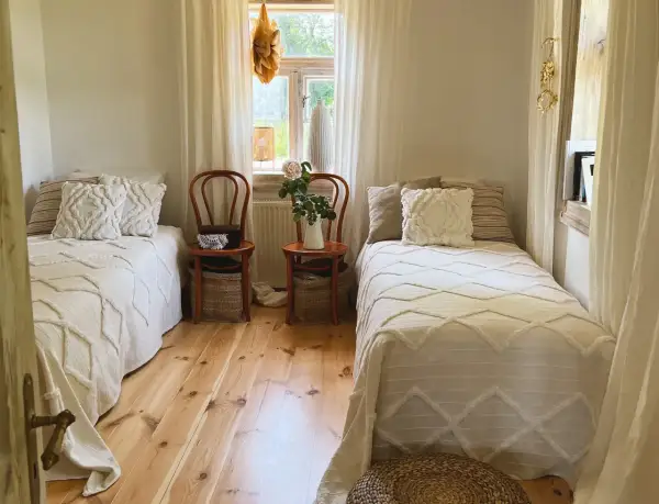 Sypialnia w stylu cottage core z dwoma łóżkami, drewnianą podłogą i oknem z firankami.