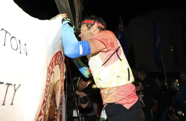 Un coureur, épuisé mais victorieux, est acclamé lors du record Hardrock 100. Des drapeaux flottent derrière lui.