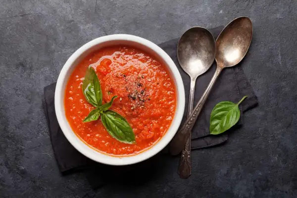 Schüssel mit bester Tomatensauce, garniert mit Basilikum und Pfeffer, daneben zwei Löffel auf dunklem Hintergrund.