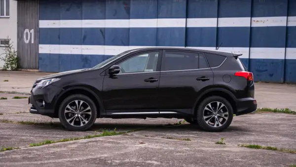 Czarny, używany Toyota RAV4 z salonu, zaparkowany przed budynkiem z niebiesko-białymi pasami.