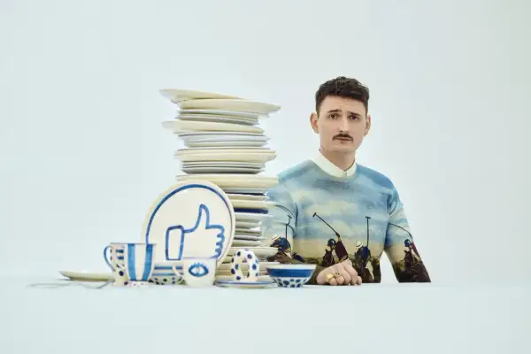 Mężczyzna z wąsikiem w swetrze z motywem polo, obok stosu talerzy i zastawy z symbolem "like". Stylizacja jak z lat dwudziestych.