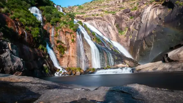 La cascada Fervenza se despliega en múltiples hilos blancos sobre rocas rojizas y verdes, desembocando en un lago sereno.