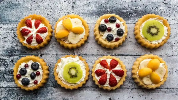 Osiem kolorowych tartaletek z owocami: truskawkami, borówkami, kiwi, bananami i morelami na kremowym nadzieniu.