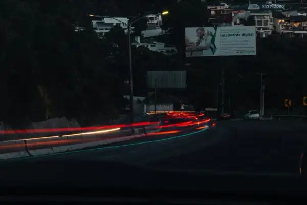 Smugi świateł samochodowych na drodze nocą, z widocznymi budynkami na wzgórzu i billboardem.