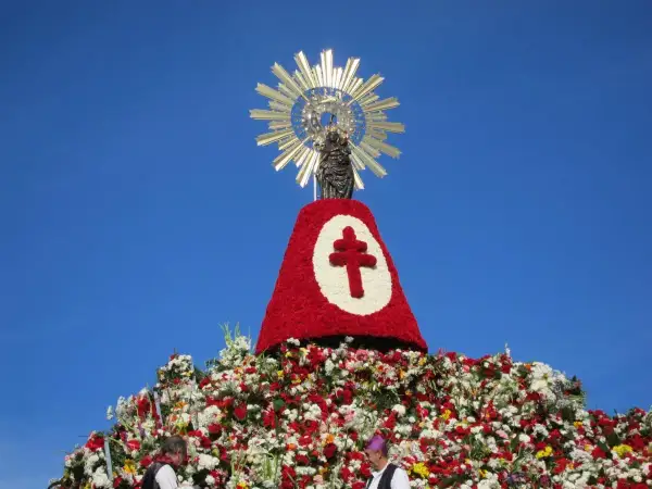 Imagen de una figura religiosa sobre un gran manto de flores rojas y blancas, con un símbolo de cruz. Se celebra una **fiesta** con **pilares** florales.
