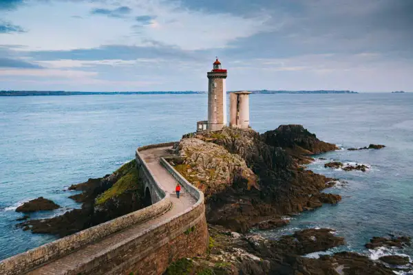 Une personne en manteau rouge marche sur la route des phares, un chemin sinueux menant à un phare sur une île rocheuse.
