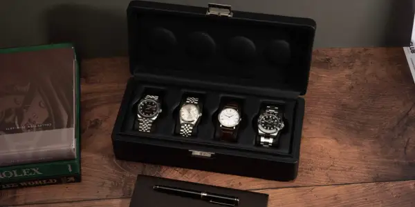 Una caja de reloj negra abierta muestra cuatro relojes de lujo. A la izquierda, libros de Rolex y un bolígrafo.