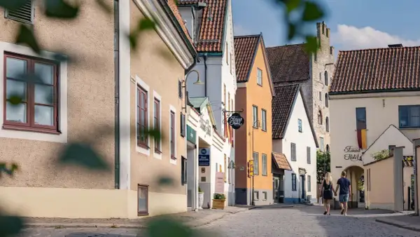 Ett par promenerar hand i hand på en kullerstensgata i Visby, förbi charmiga hus och skyltar som indikerar B&B Gotland och Strand Hotel.