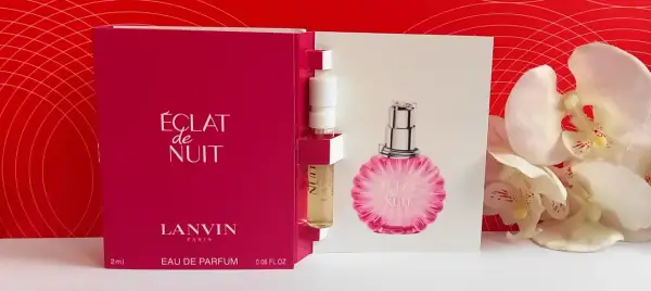 Próbka perfum Lanvin Éclat de Nuit, z buteleczką i kartonikiem z wizerunkiem flakonu, na tle czerwonych wzorów i białych kwiatów.