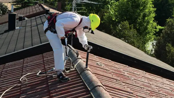 Pracownik w białym kombinezonie i żółtym kasku usuwa stare pokrycie dachowe, prawdopodobnie eternit, z czerwonego dachu.