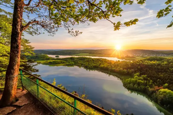 Vue panoramique depuis le belvédère Pic de l'Aigle au coucher du soleil, avec un lac reflétant le ciel et des collines verdoyantes.