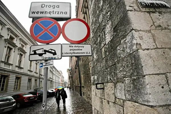 Znaki drogowe informują o zakazie zatrzymywania się i postoju na drodze wewnętrznej. W tle widać ulicę z samochodami i przechodniów.